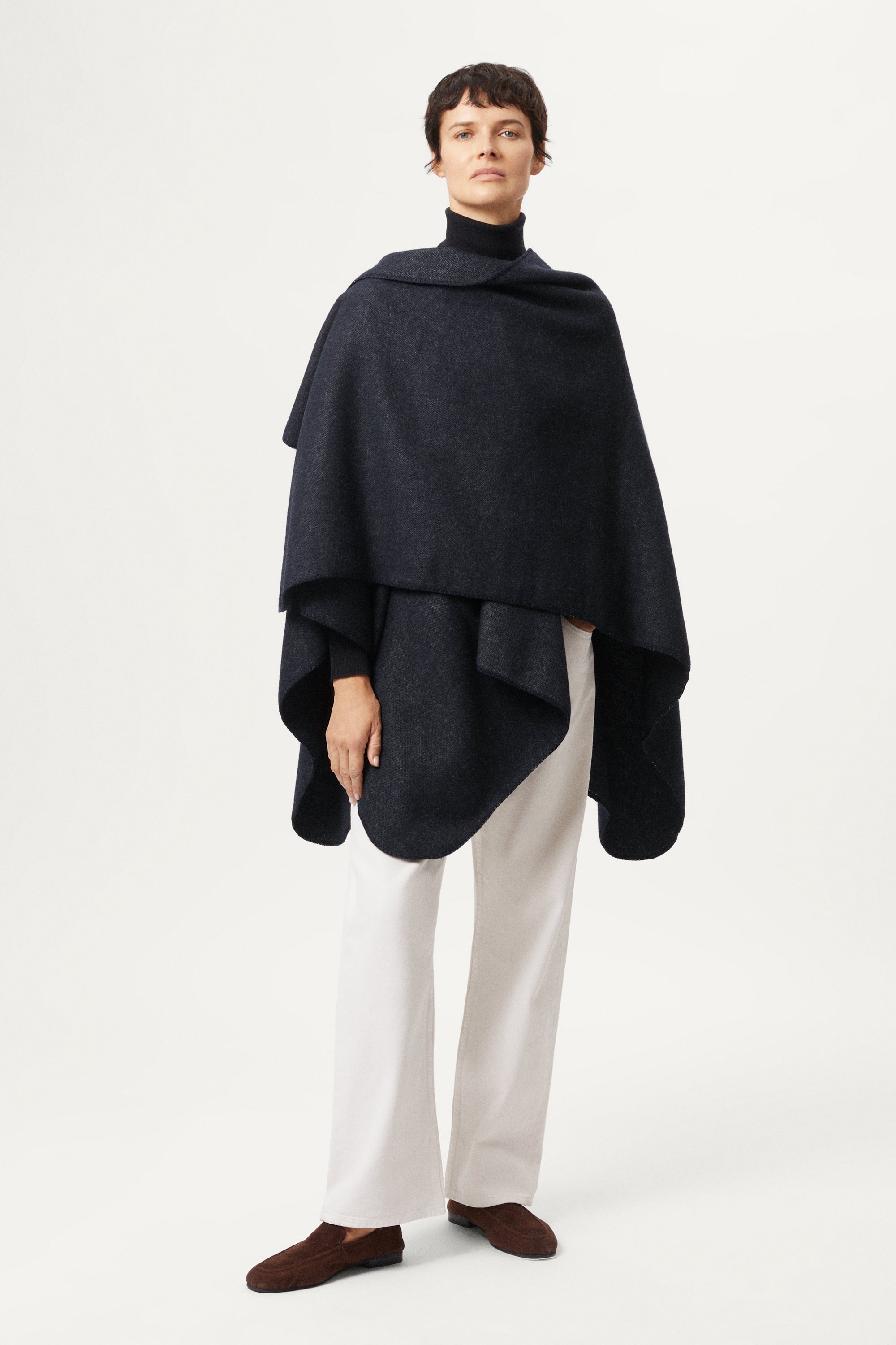 woman wearing a blue navy poncho over a turtleneck and white pants on a white background