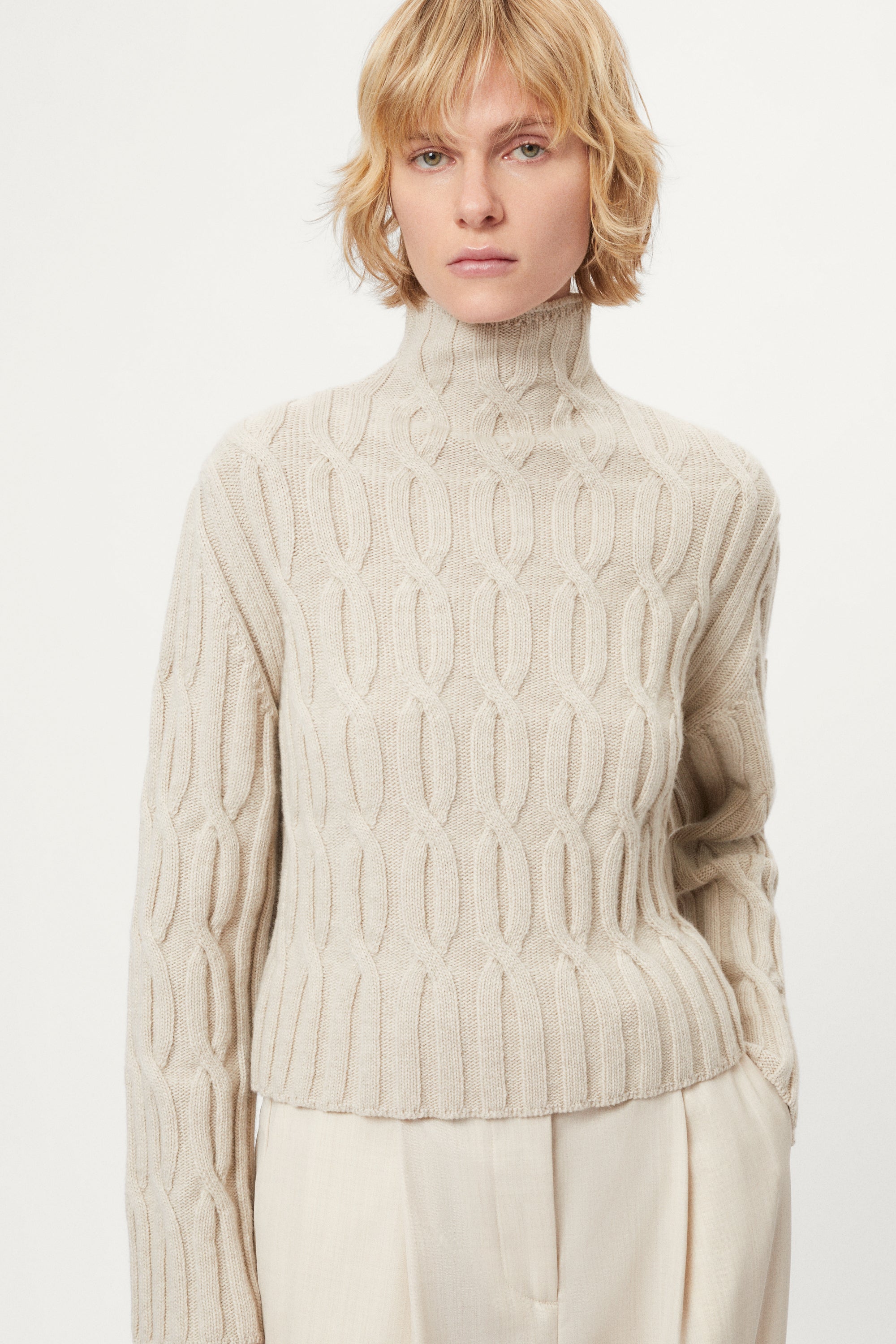 woman wearing a woolen  beige cable knit turtleneck sweater on a white background